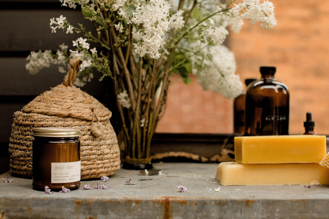 A Field + Bee candle beside a woven bee skep, white blossoms, amber bottles and stacked beeswax on the garden potting table