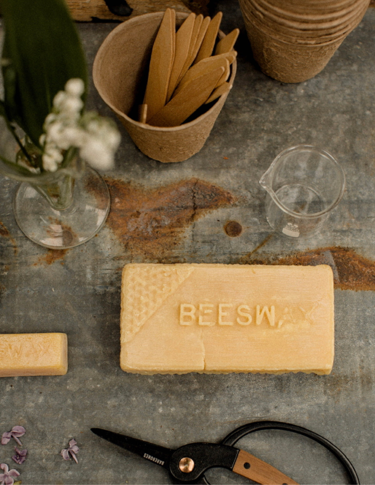 Golden block of beeswax on a weathered zinc worktable beside a glass beaker, flowers, wooden tools and scissors.